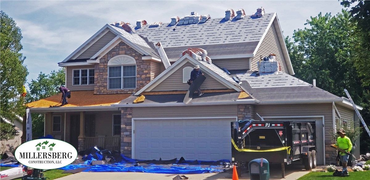 Roofers working on a house with removed shingles. A dumpster trailer is parked in the driveway.