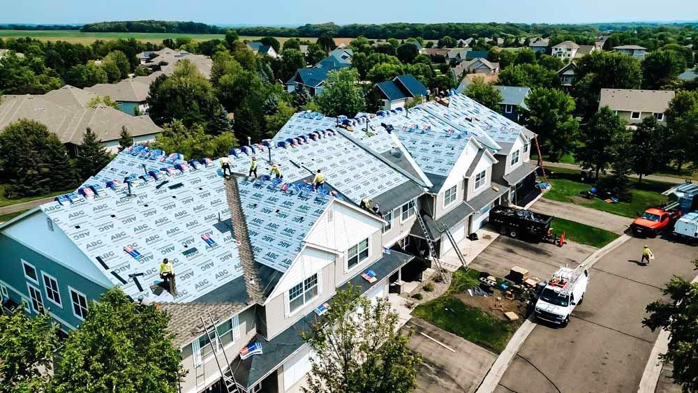 Roofers working on multiple houses in a residential area, blue tarp visible on the roofs.