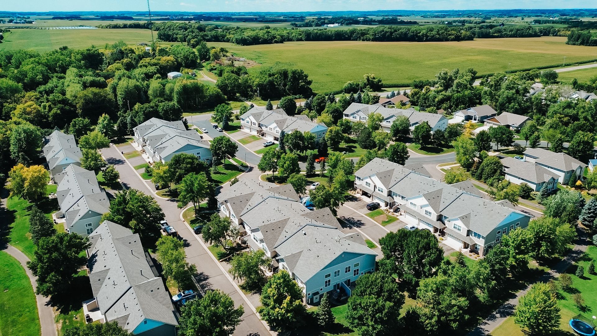 Aerial view of a suburban neighborhood with rows of houses, trees, and a field in the background under a blue sky.