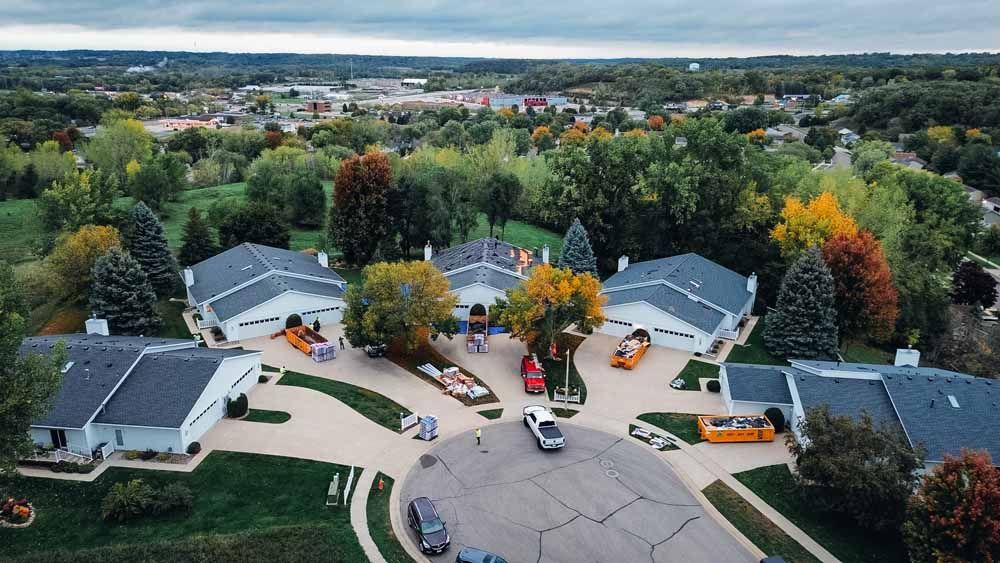 Aerial view of houses on a cul-de-sac. Cars and orange construction vehicles are present. Fall foliage surrounds the neighborhood.