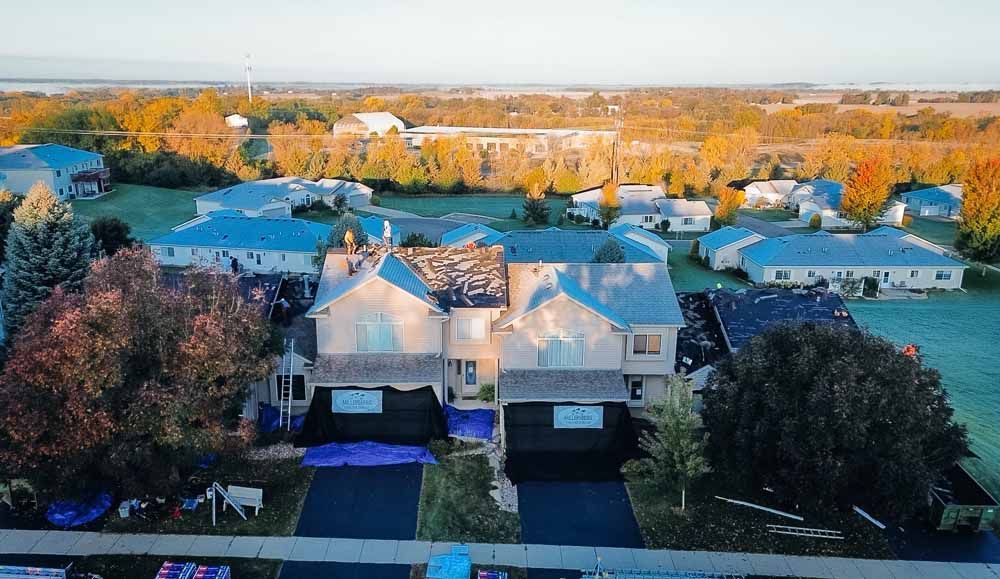 Aerial view of a damaged house with tarps on the roof and driveways, surrounded by other houses and trees.