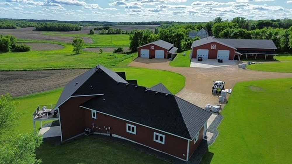 Red house with black roof, three red garages, and green fields in a rural setting on a sunny day.