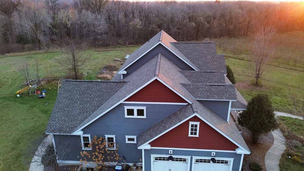 Aerial view of a blue house with a red gable roof, at sunset. Green lawn and trees surround the home.