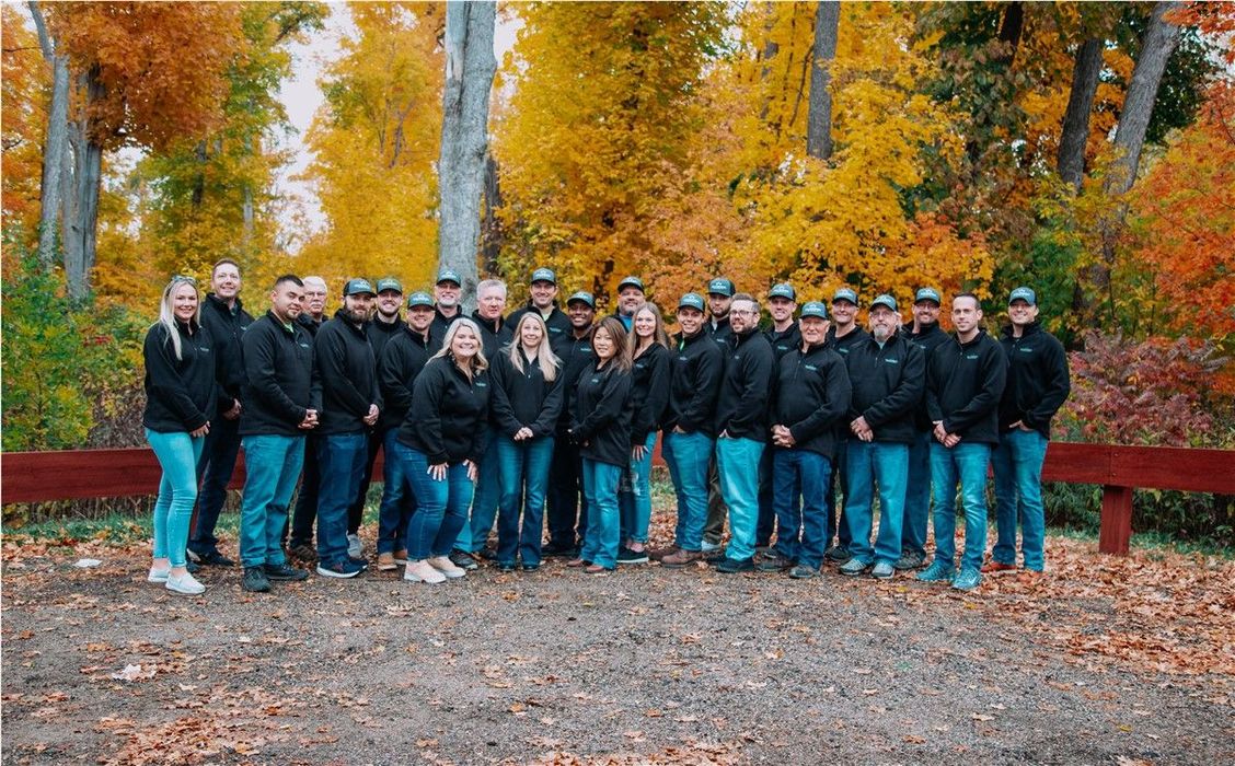 Group of people wearing black jackets and blue jeans, standing in front of fall foliage.
