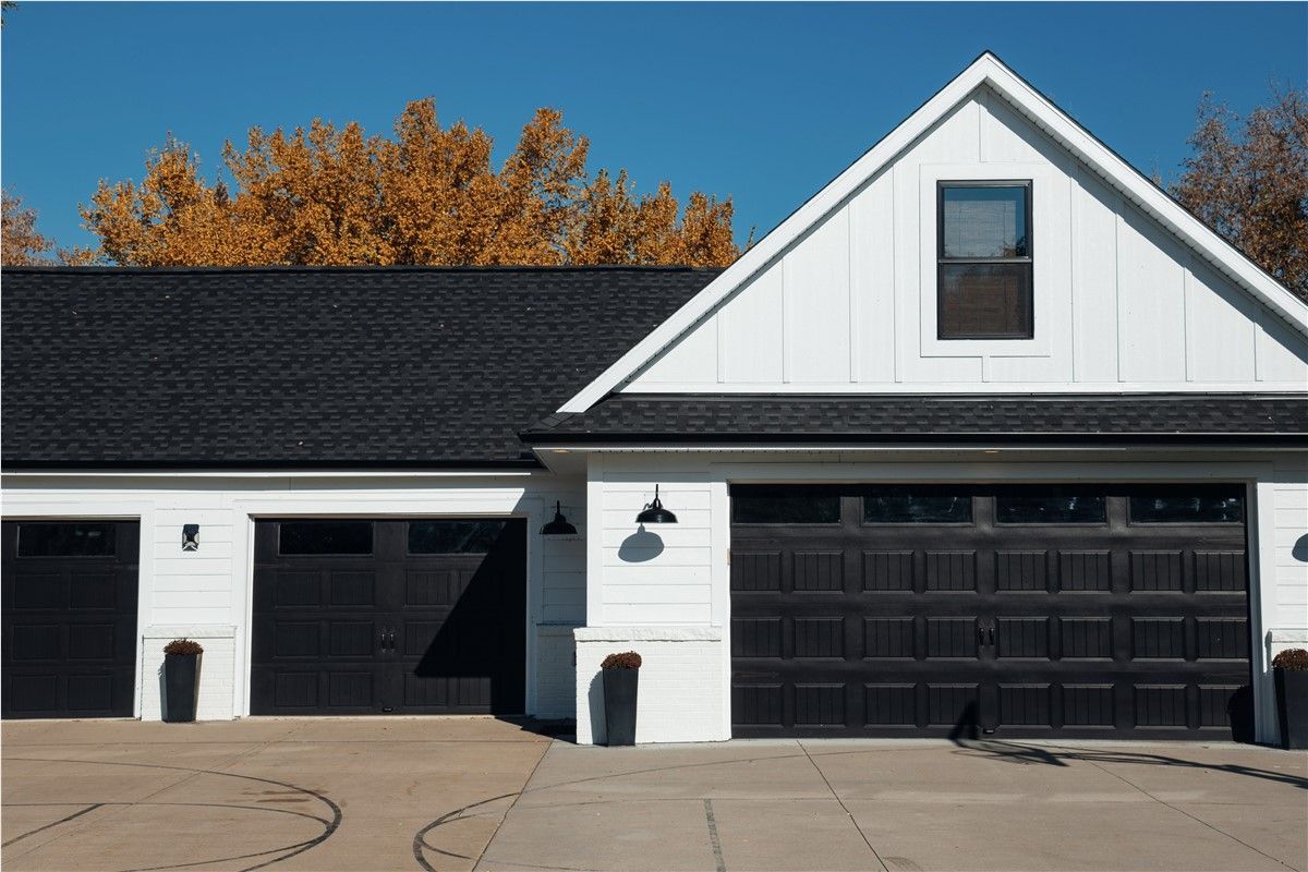 White farmhouse-style garage with three black garage doors, black roof, and autumn trees.