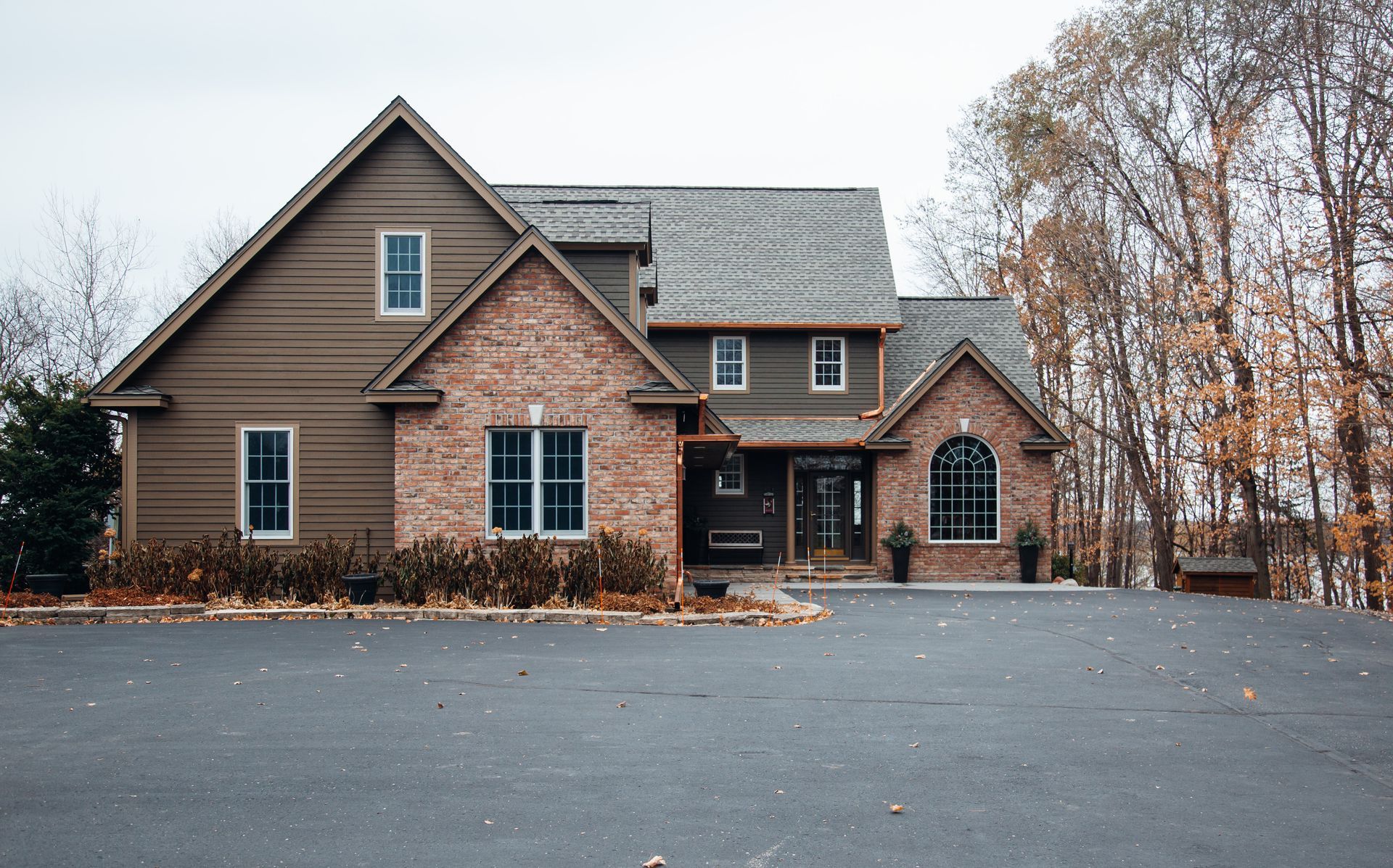 Two-story house with brown siding, brick accents, and a dark asphalt driveway. Trees in background.