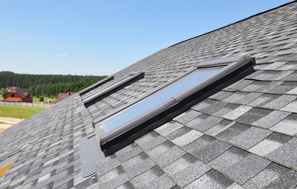Three rectangular skylights on a gray shingle roof under a blue sky.