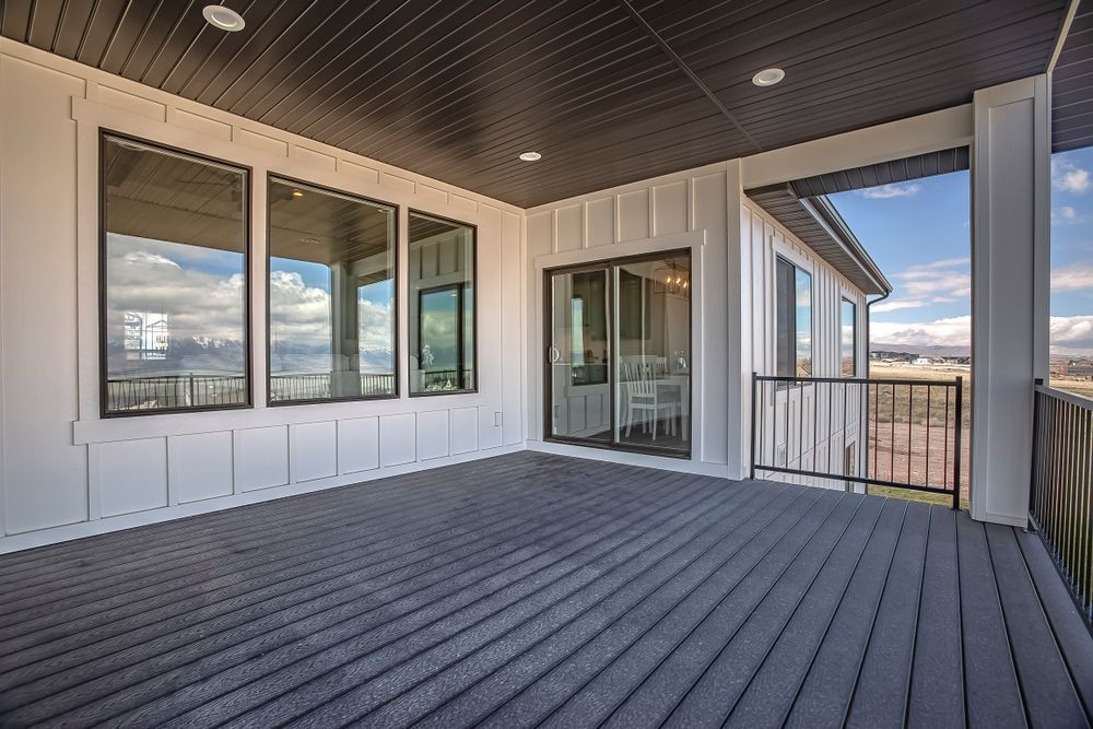 Covered outdoor deck with dark gray floorboards, white siding, black-framed windows, and railing, overlooking a scenic view.