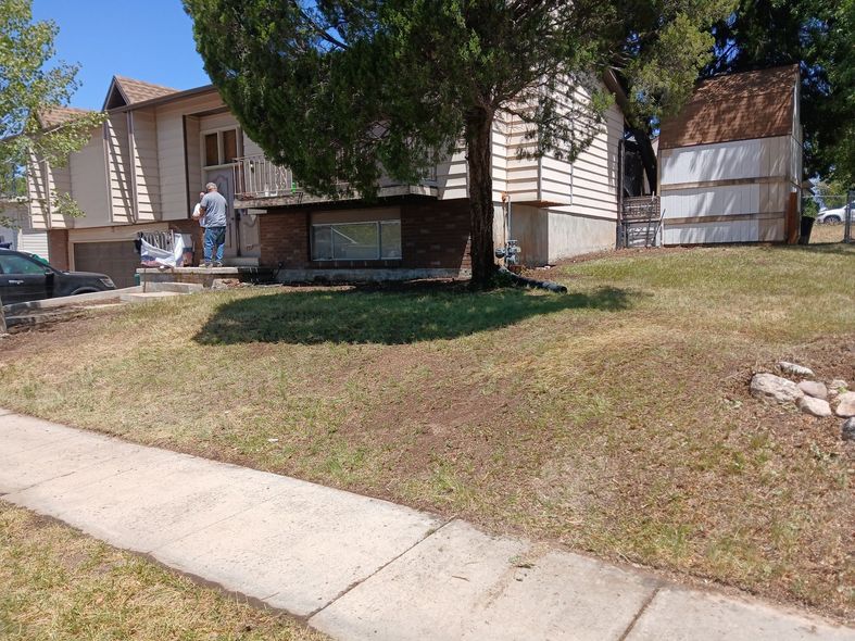 A person stands outside a two-story house with a detached shed on a sunny day. The lawn is brown and dry.