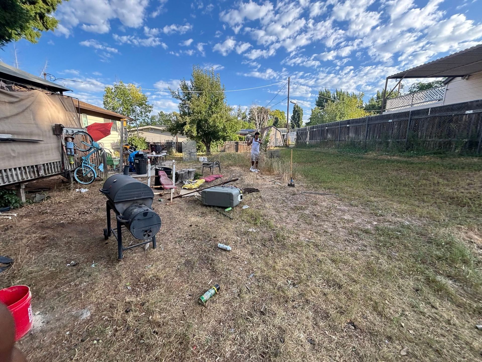 Backyard with a barbecue, debris, and a person standing near a fence under a partly cloudy sky.