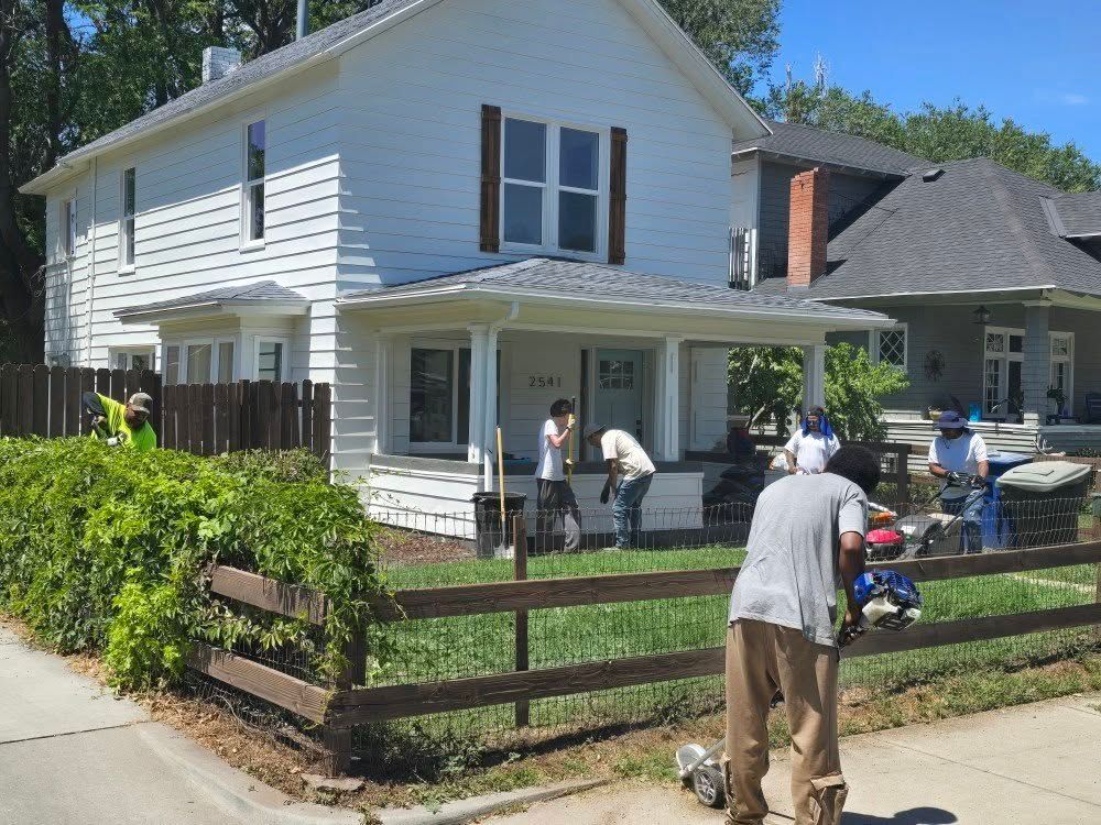 Workers landscaping a white house with a porch and a wooden fence, using tools on a sunny day.