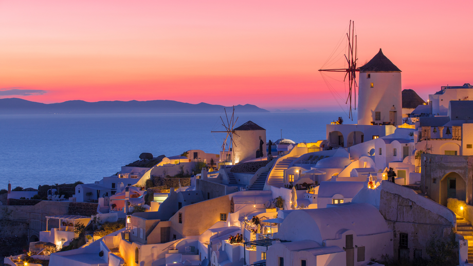 Sunset over a cliffside Greek village with white buildings, windmills, and the sea beyond