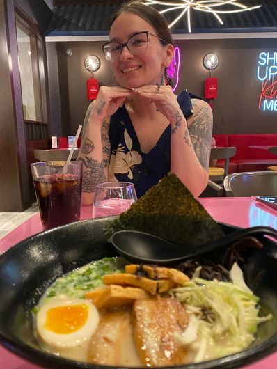 Woman smiling at a restaurant table with ramen, drinks, and a neon-lit background
