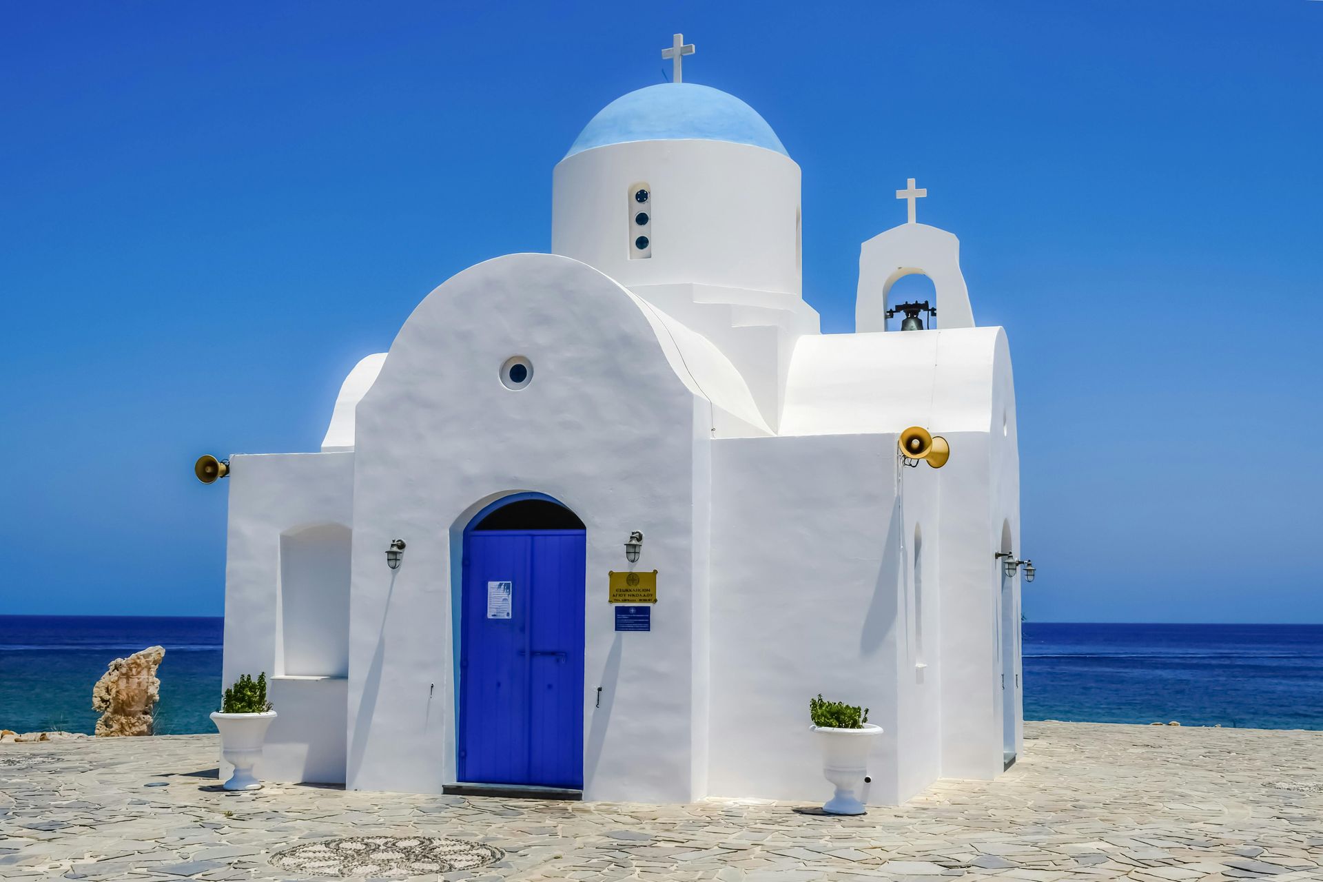 White seaside chapel with blue doors and domes against a bright blue sky and ocean.
