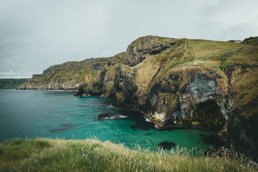 Rocky coastal cliffs above a turquoise cove with grassy foreground and calm sea