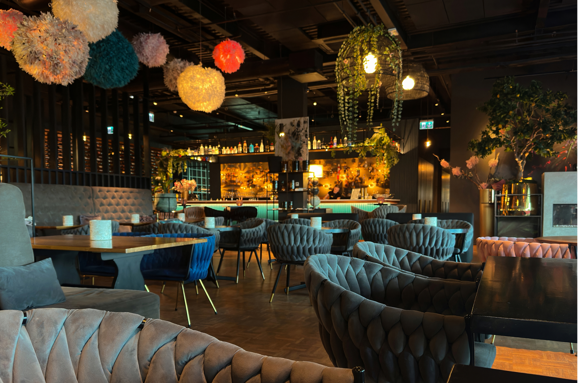 Restaurant interior with plush seating, bar in background, decorated with hanging lights and plants.