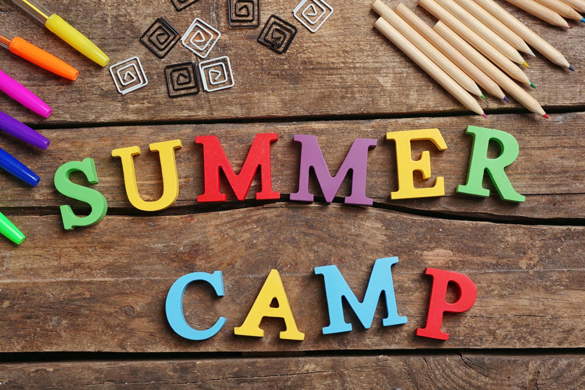 The word summer camp is written in colorful wooden letters on a wooden table.