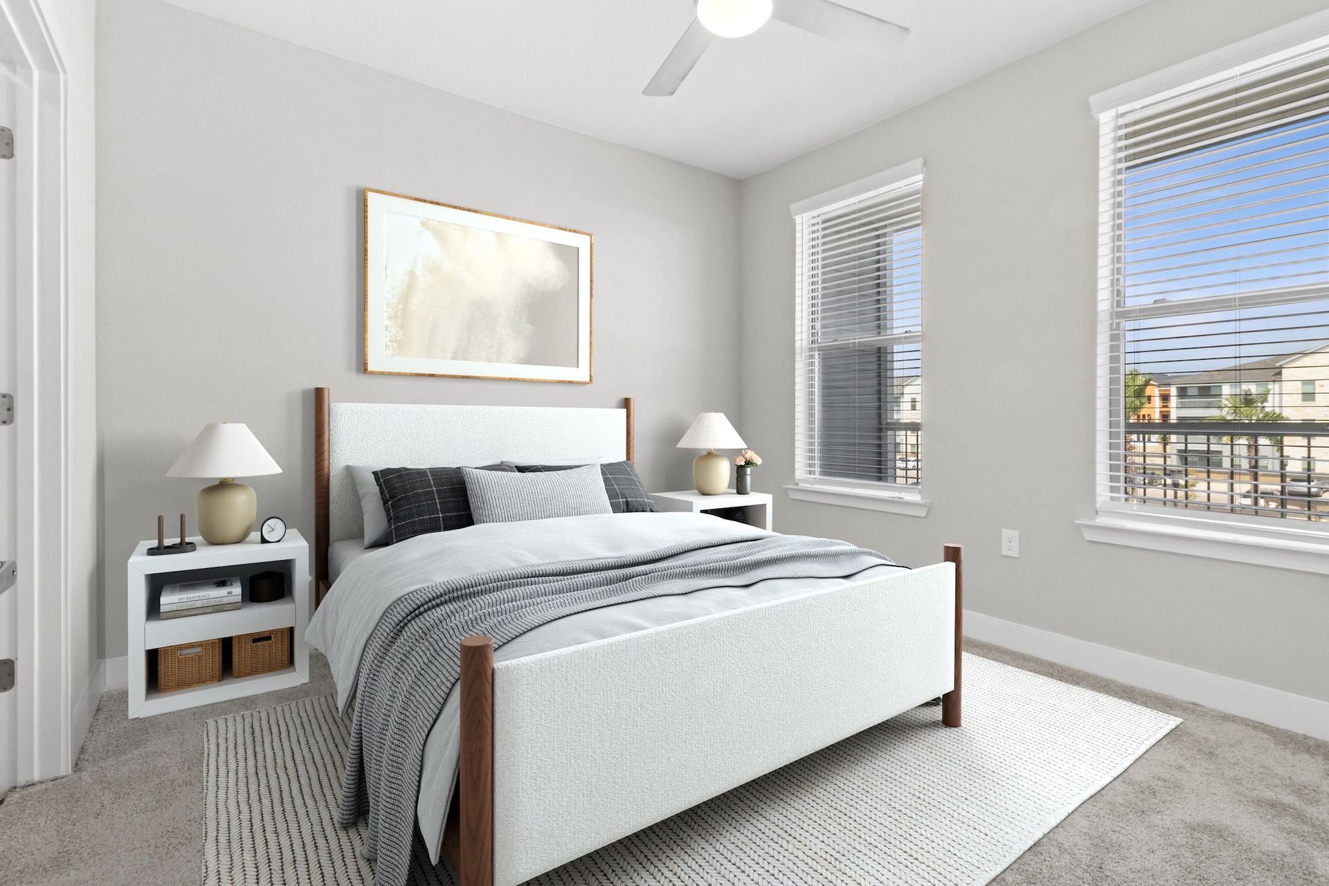 Bedroom with a white bed, nightstands, and a framed artwork. Two windows with blinds provide natural light.