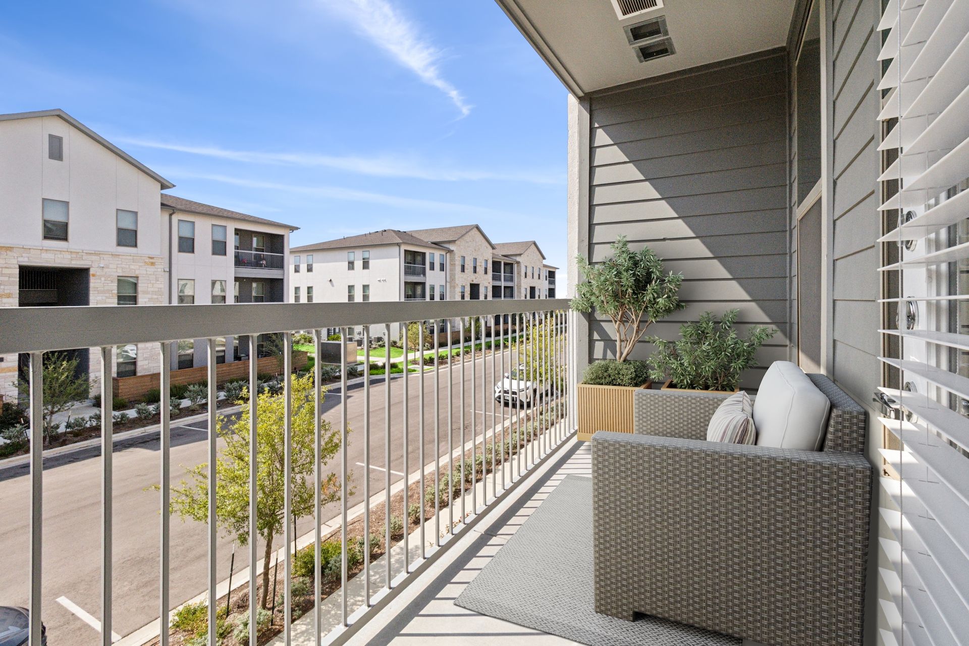 Balcony with wicker chair, rug, and plants overlooking a street and buildings on a sunny day.