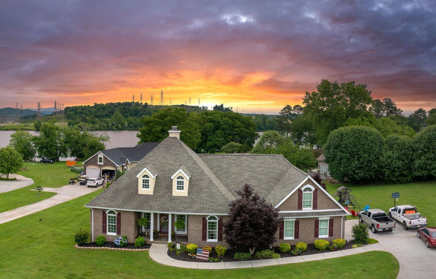 A single-story brick house with a gray roof and dormer windows at sunset, overlooking a lake with trees and parked cars.
