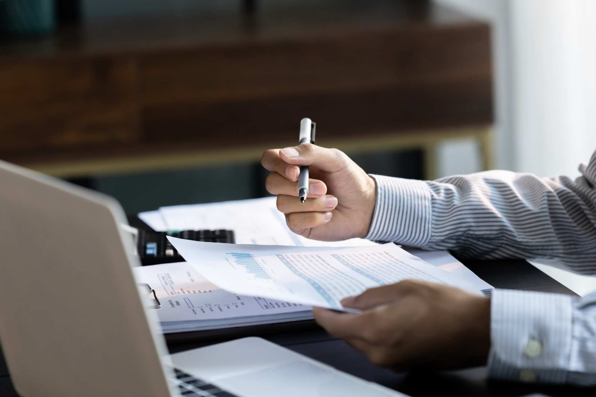 Hands holding a pen and financial documents at a desk with a laptop, focusing on work tasks.