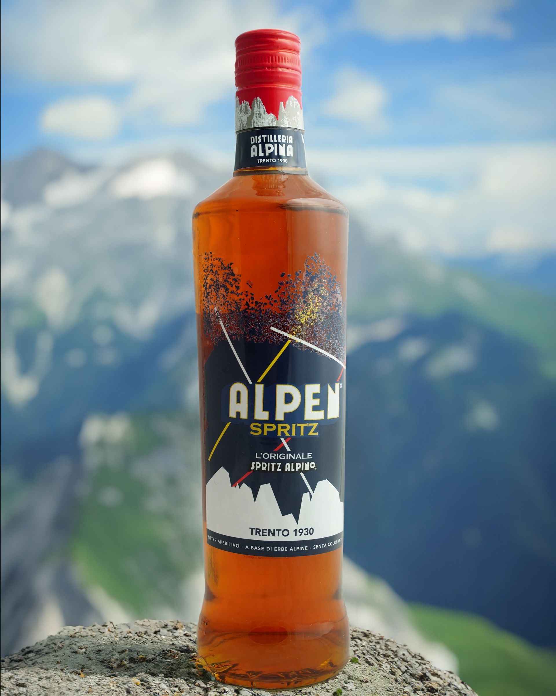 Bottiglia di Alpen Spritz con etichetta, sullo sfondo di montagne. Colori rosso, nero e bianco.
