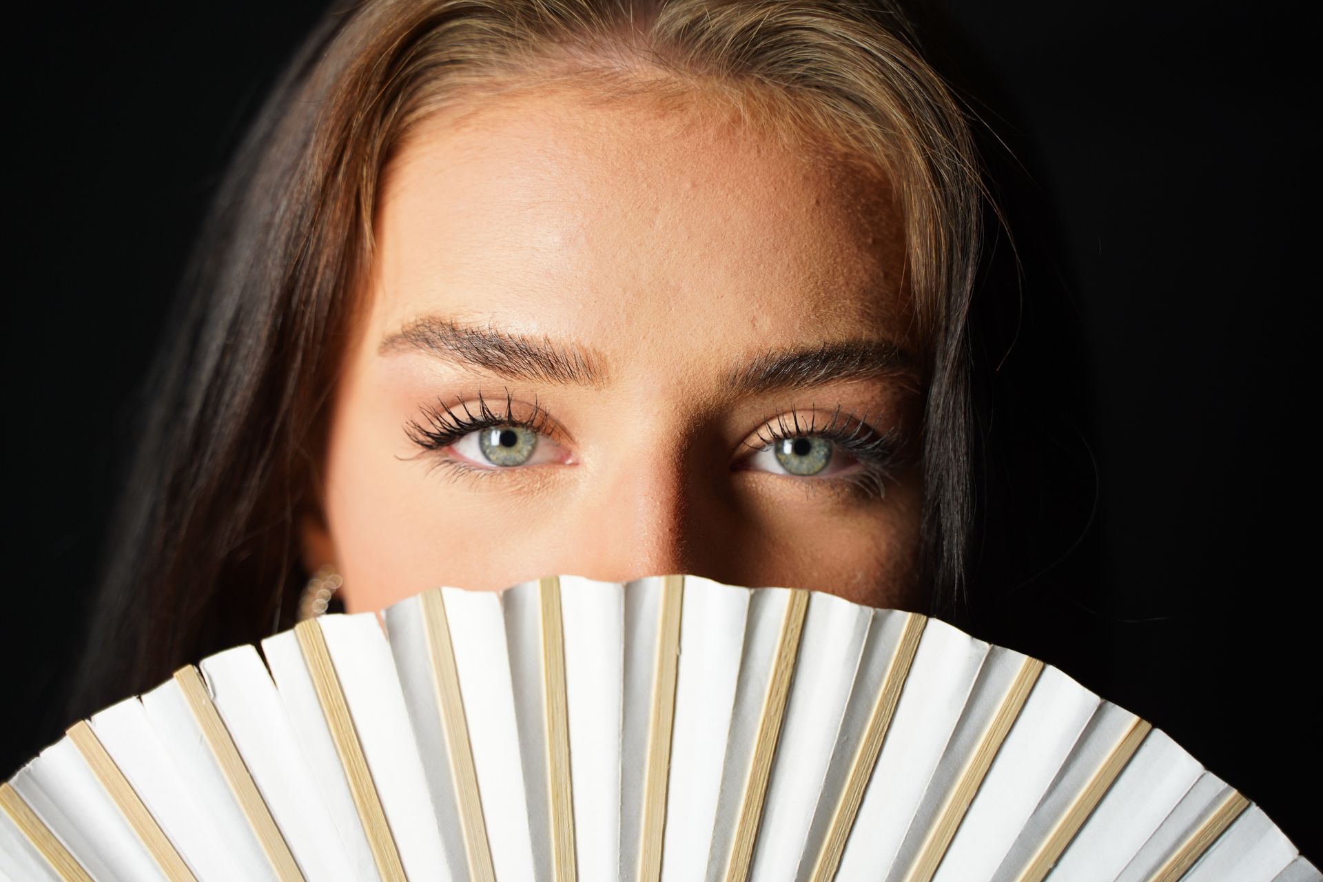 Donna con occhi verdi, che tiene in mano un ventaglio bianco e oro, e guarda verso la telecamera.
