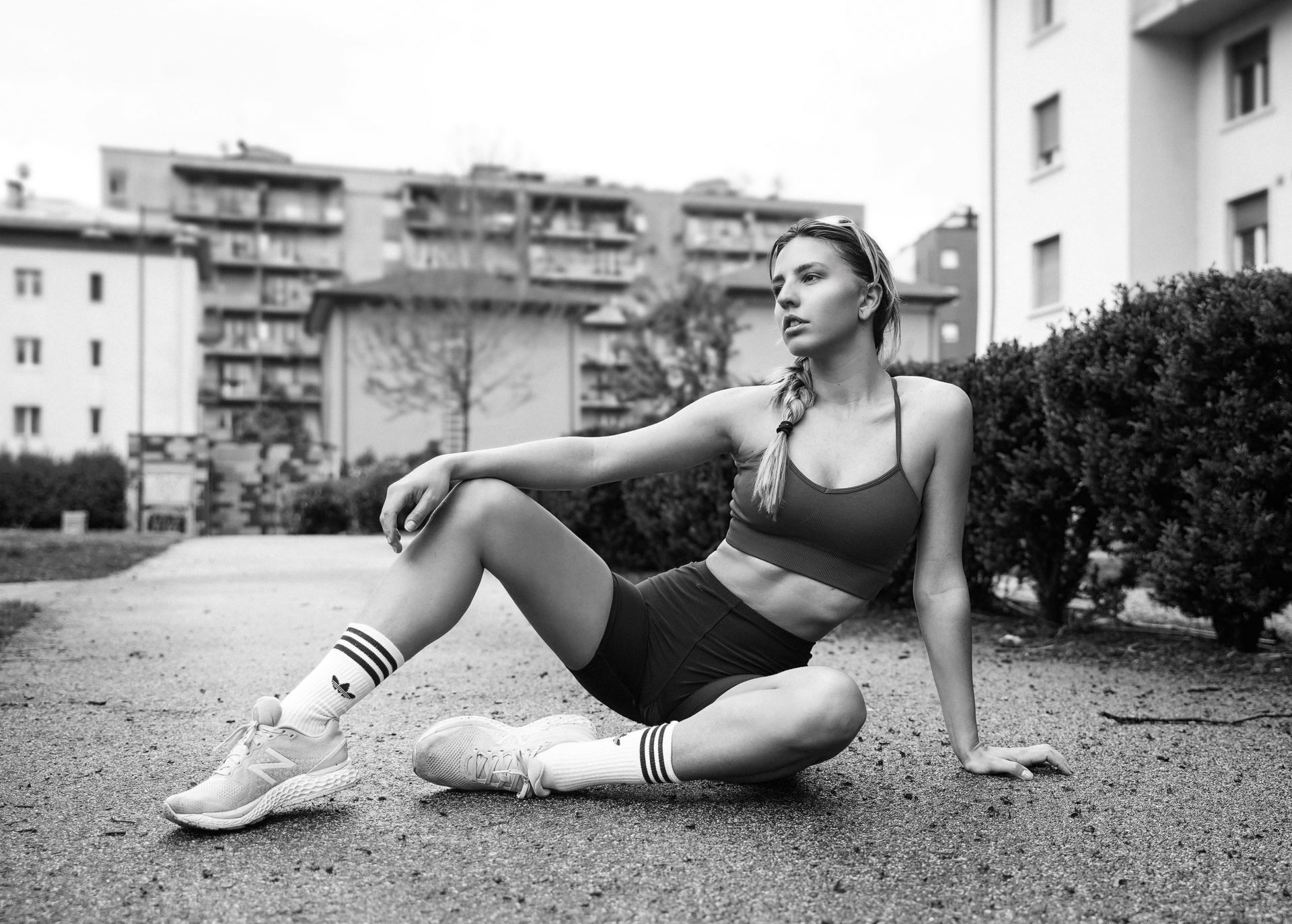 Donna in abiti sportivi, seduta per terra all'aperto. Guarda di lato con un'espressione seria.