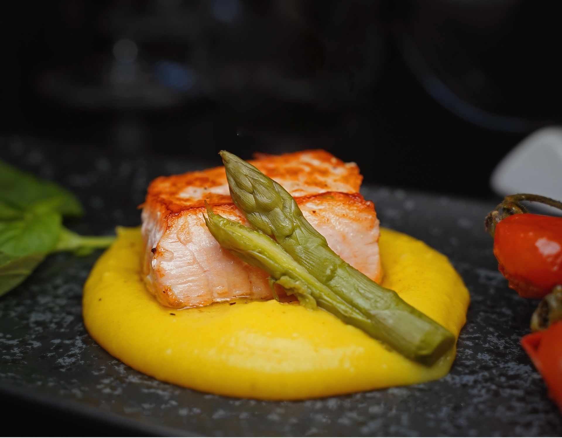 Salmone scottato su un letto di purea gialla, con asparagi e pomodori, su un piatto scuro.