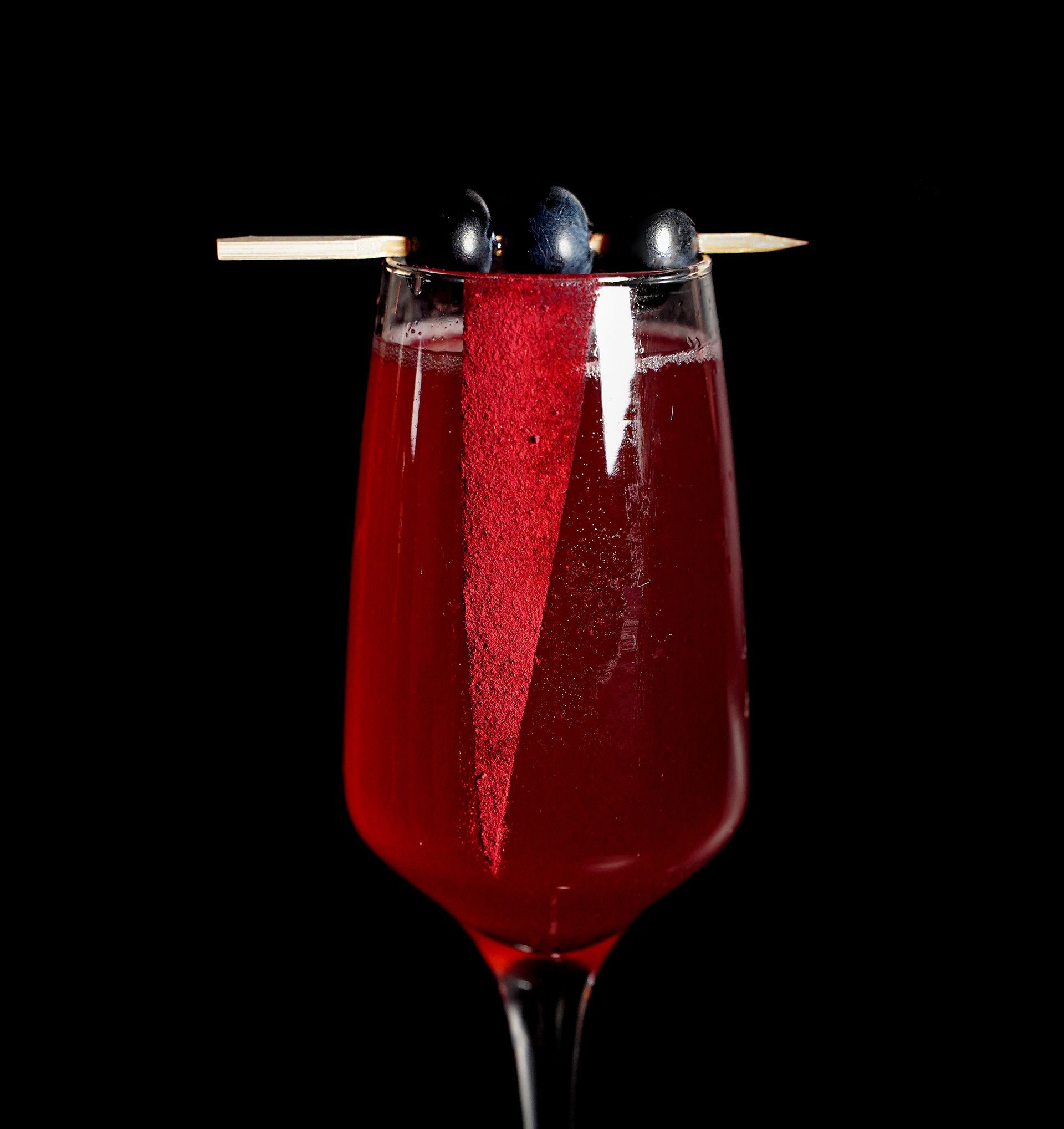Cocktail rosso in un bicchiere con guarnizione bordata di zucchero rosso e tre mirtilli infilzati su uno spiedino.