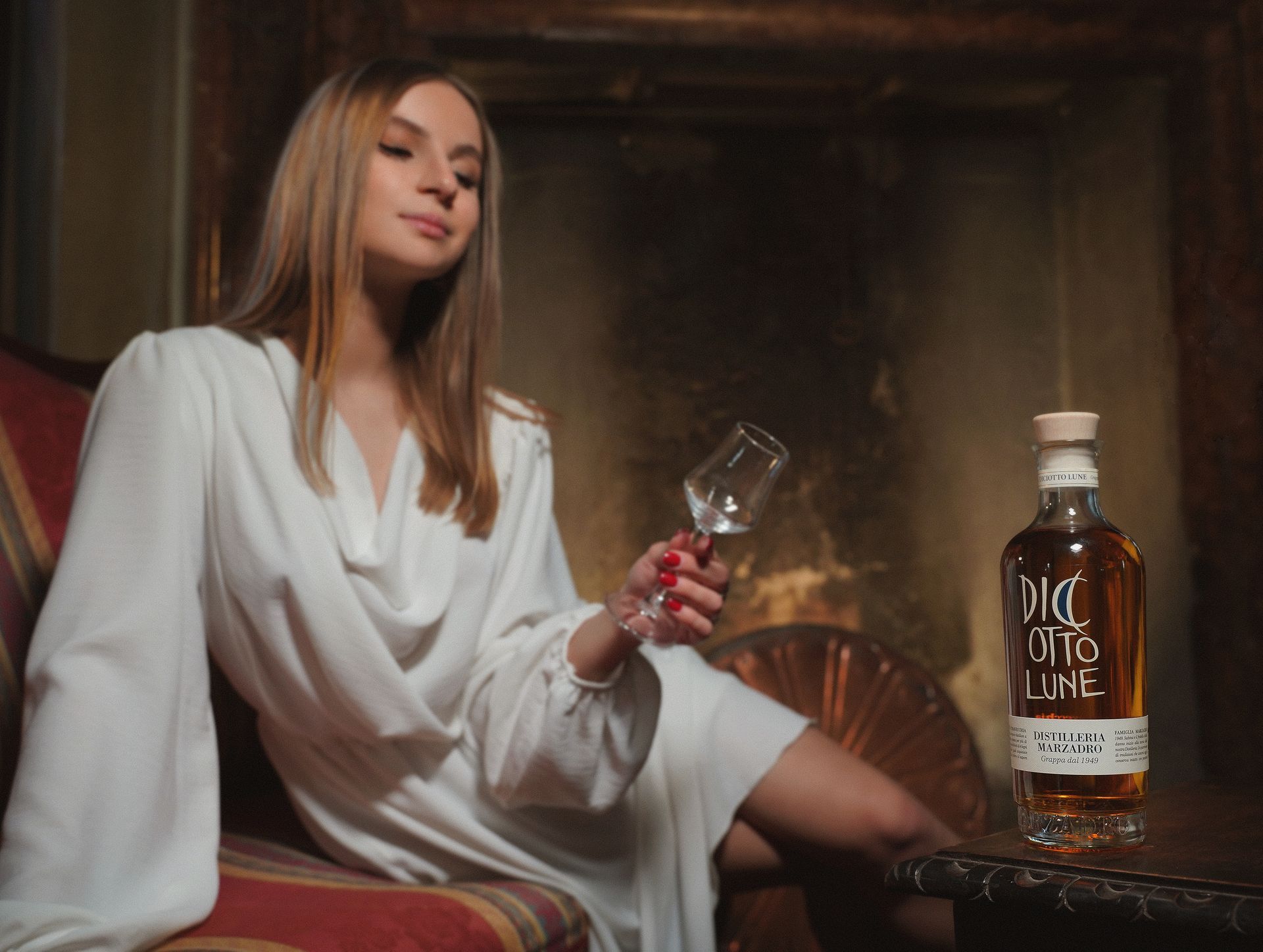 Donna in abito bianco che tiene in mano un bicchiere e una bottiglia di liquore, seduta accanto al camino.