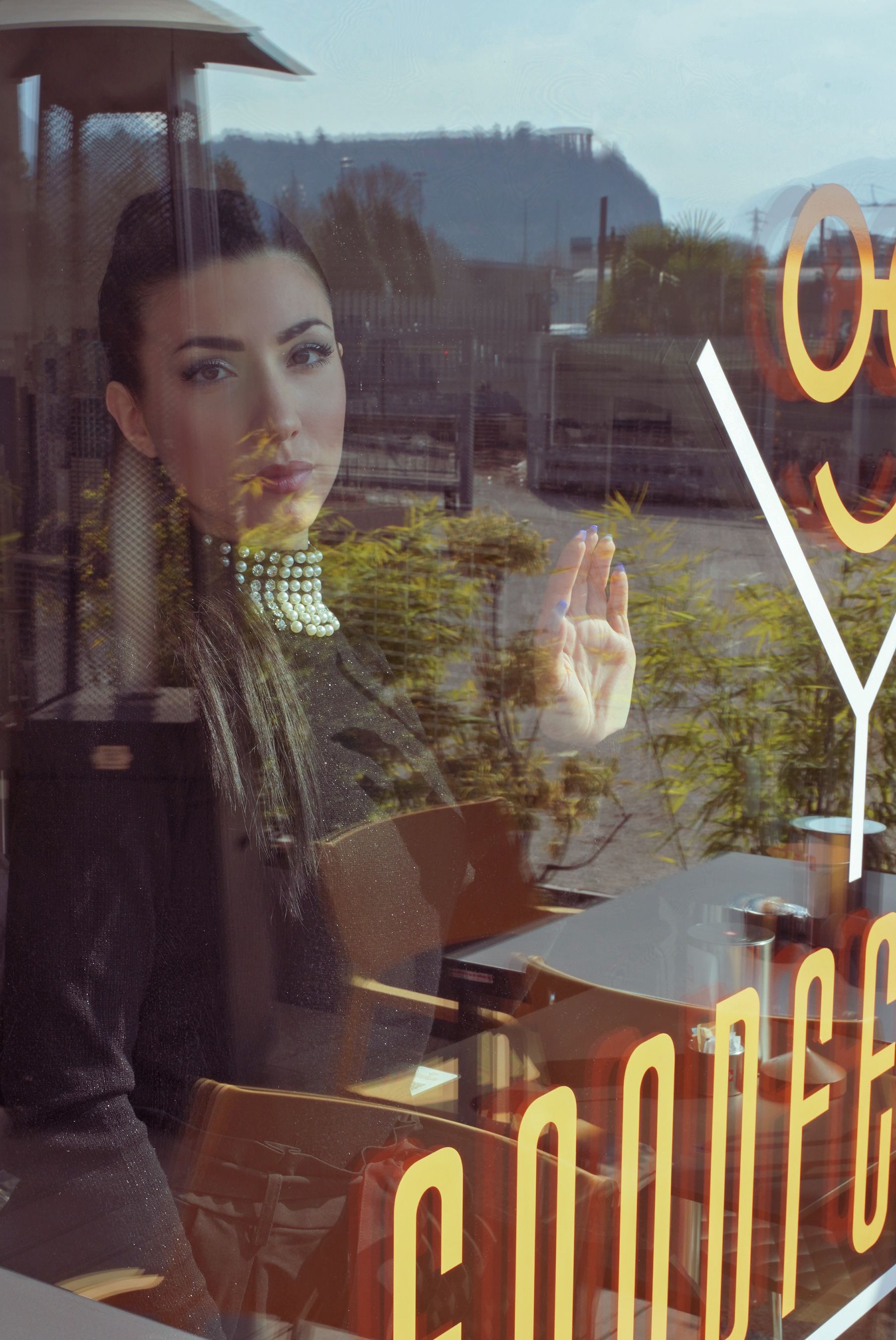 Donna al bar, guarda fuori dalla finestra, tocca il vetro. È visibile la scritta 