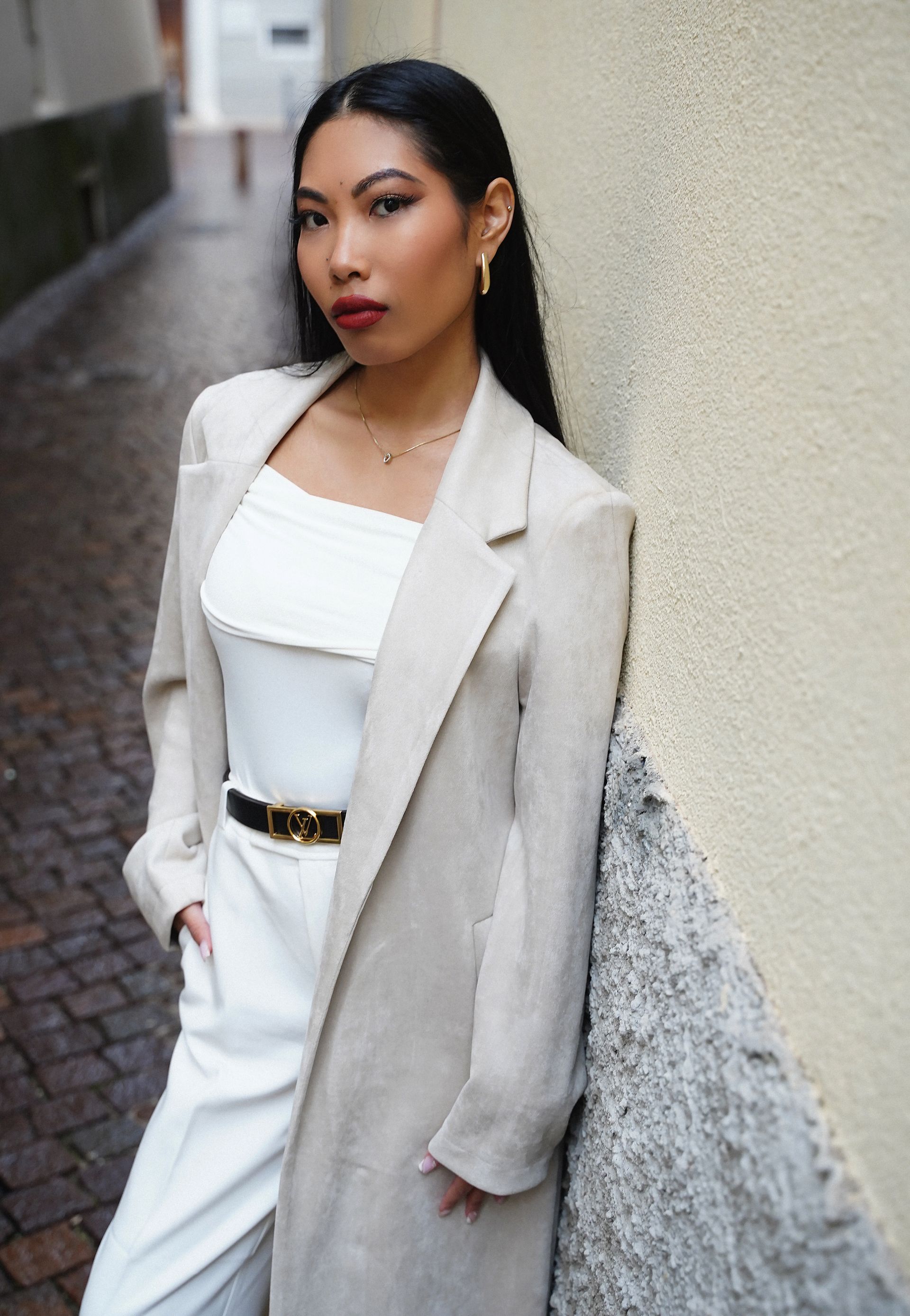 Una donna con giacca beige e abito bianco è appoggiata a un muro in un vicolo e guarda la telecamera.