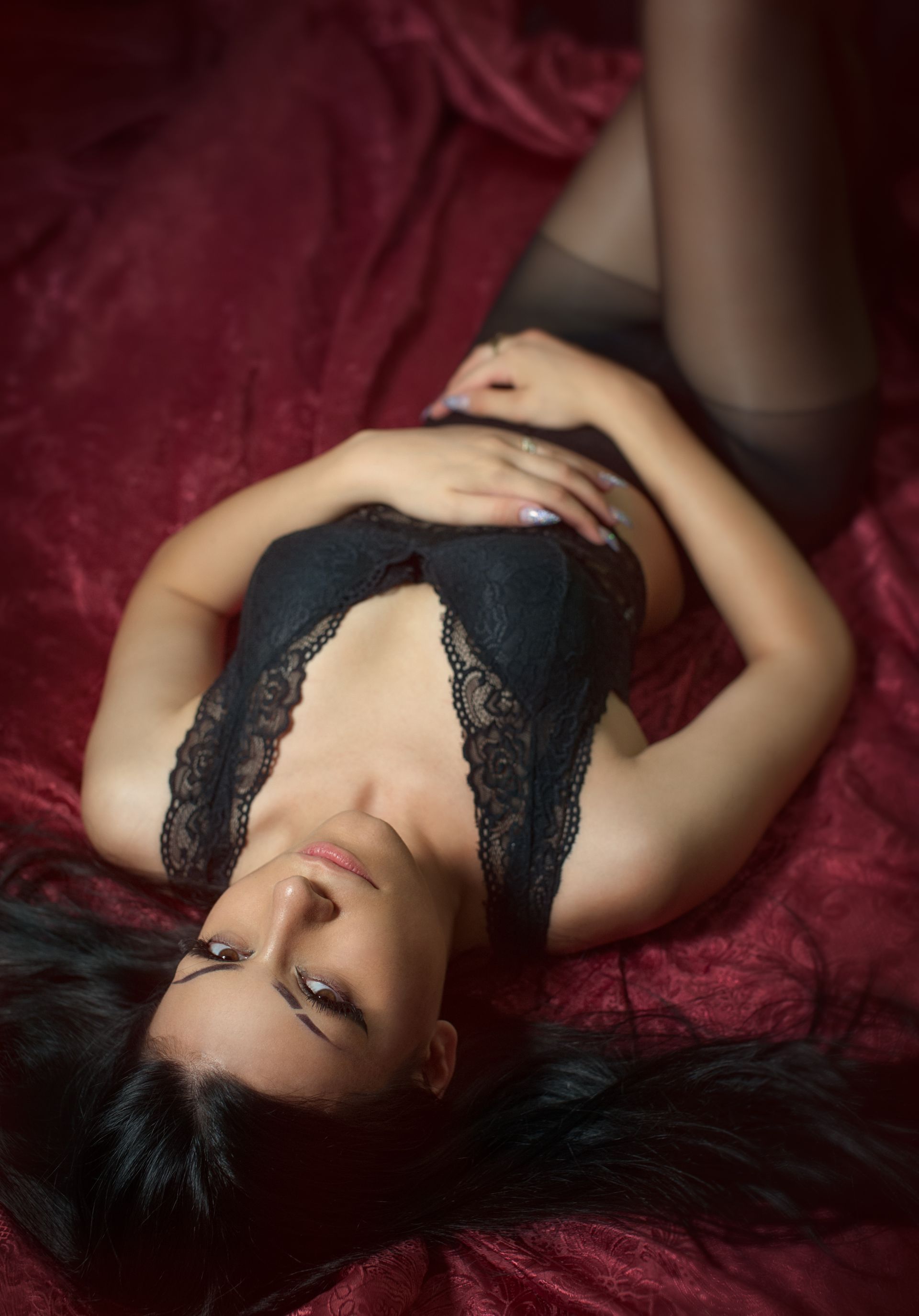 Una donna in lingerie nera e collant è sdraiata su un tessuto bordeaux e guarda l'osservatore.