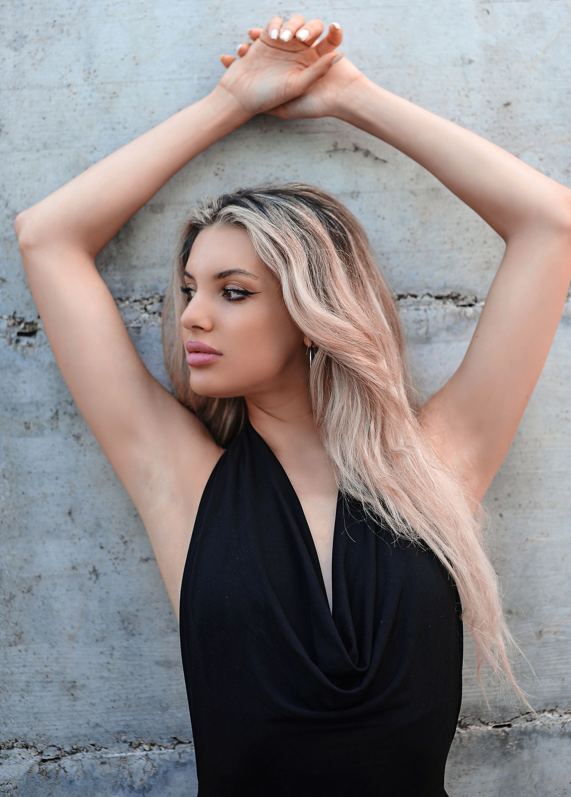 Donna in abito nero, mani sopra la testa, appoggiata a un muro di cemento grigio.