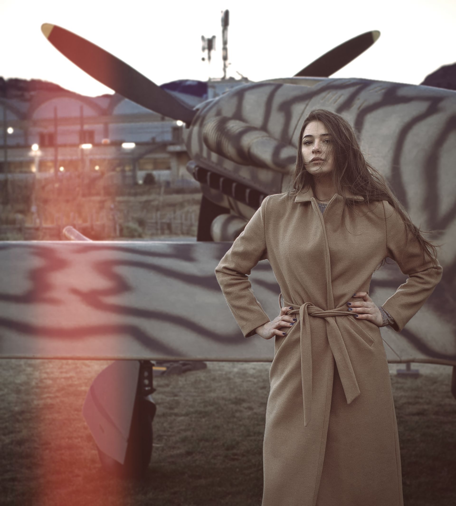 Una donna con un cappotto color marrone chiaro sta in piedi con sicurezza davanti a un aereo a elica mimetizzato al tramonto.
