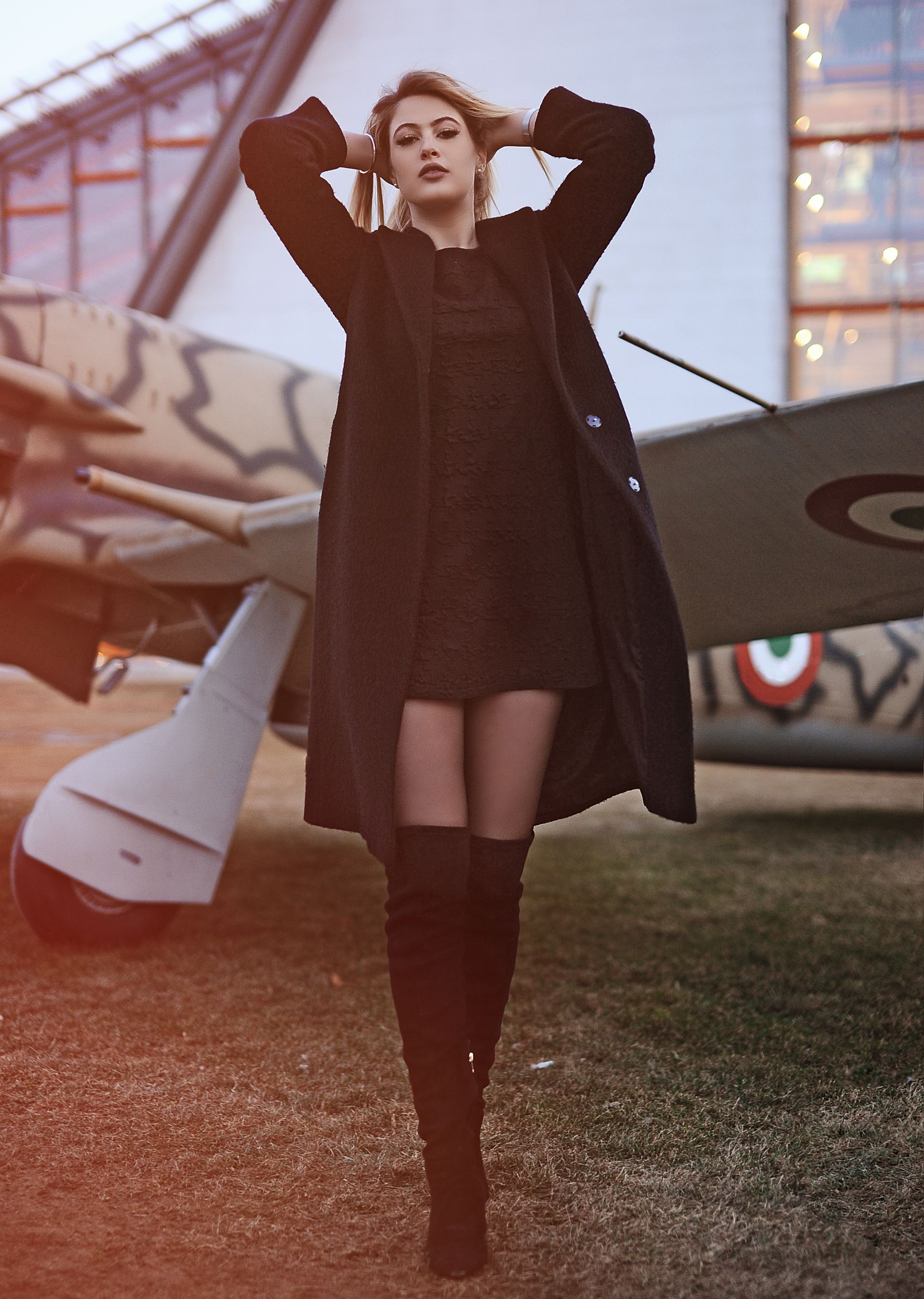 Una donna con cappotto nero e stivali, mani sulla testa, è in piedi vicino a un aereo d'epoca.