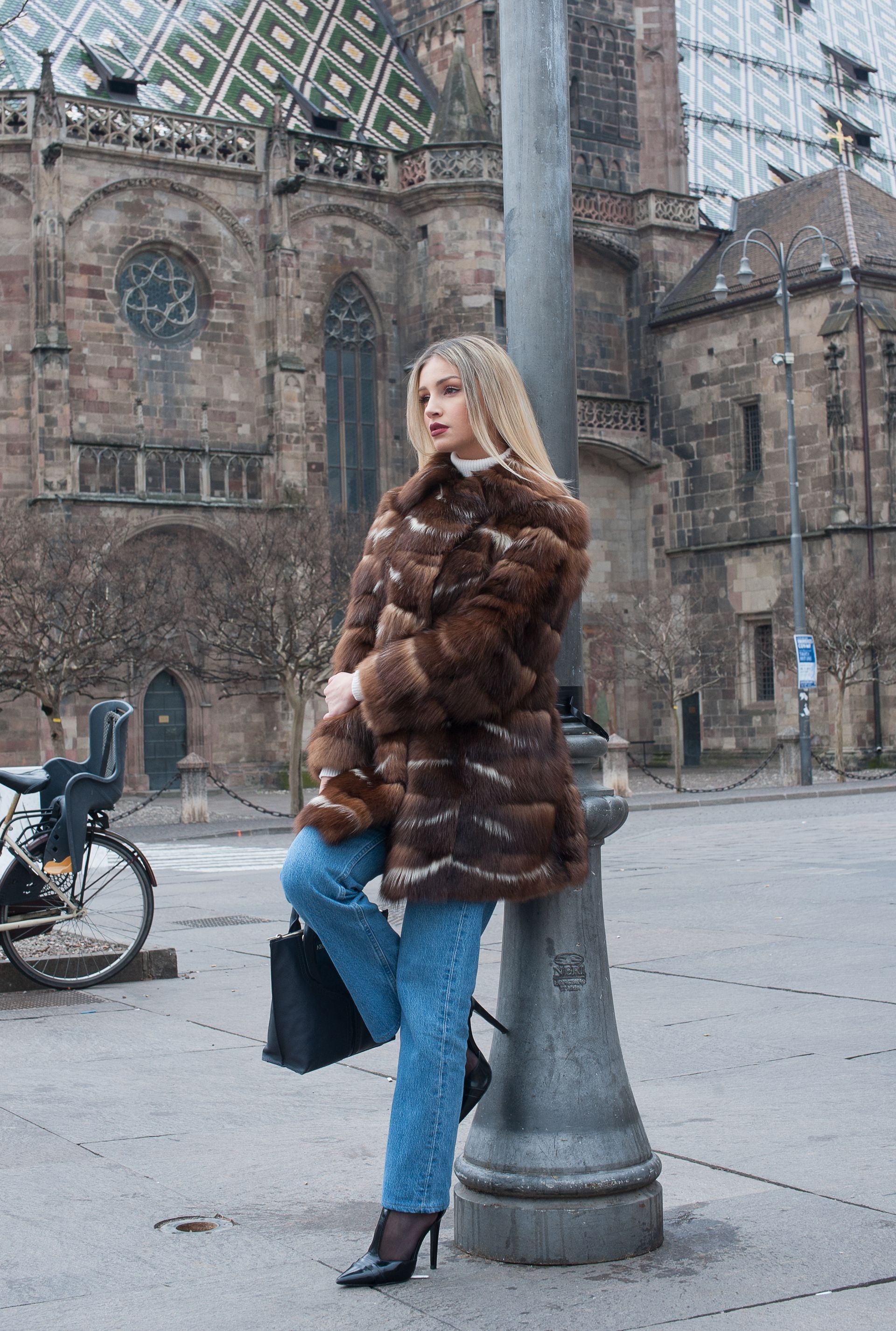 Una donna con un cappotto di pelliccia marrone e jeans si appoggia a un lampione di fronte a una cattedrale.