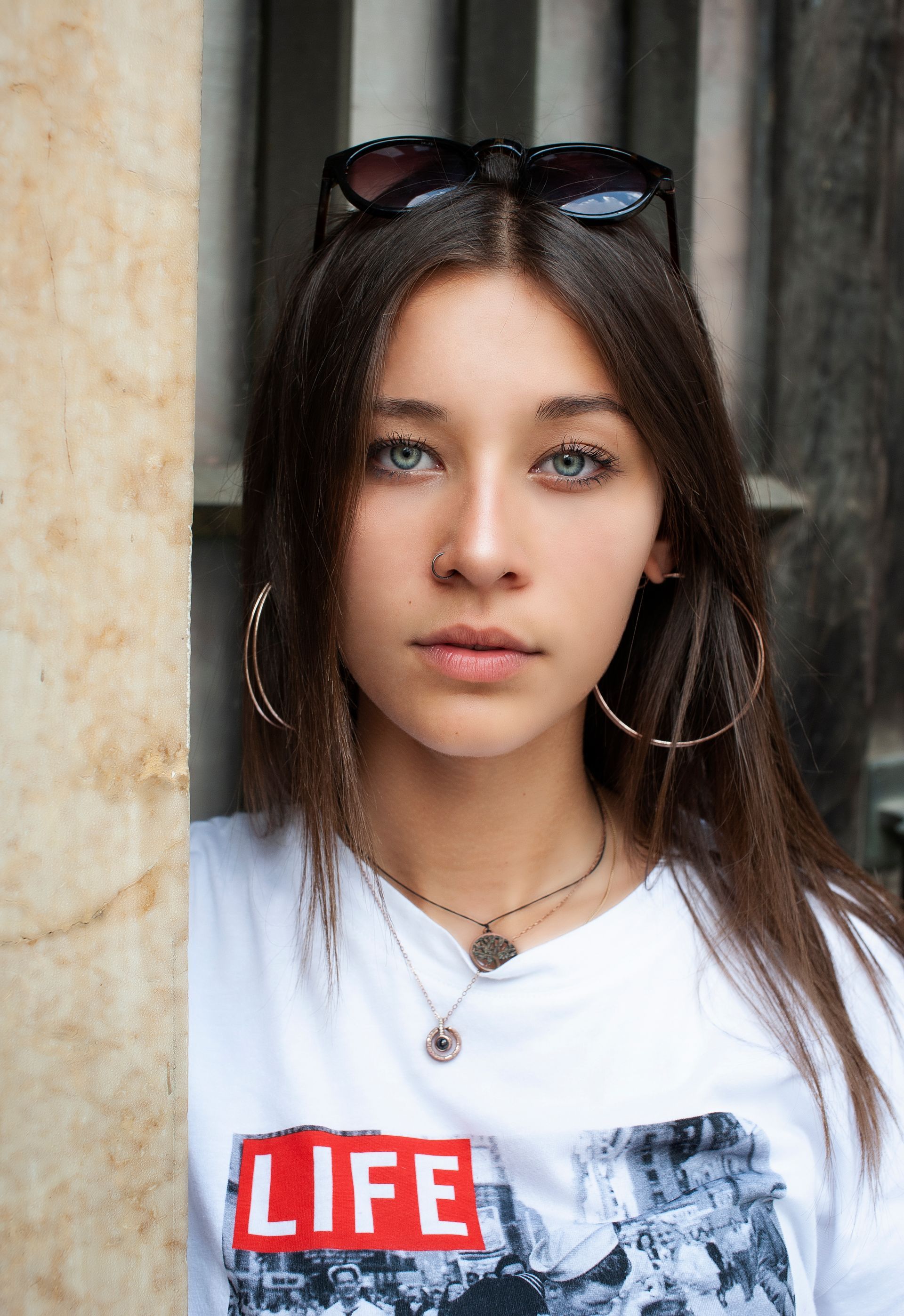 Giovane donna con orecchini a cerchio, occhiali da sole in testa, maglietta bianca, occhi azzurri.