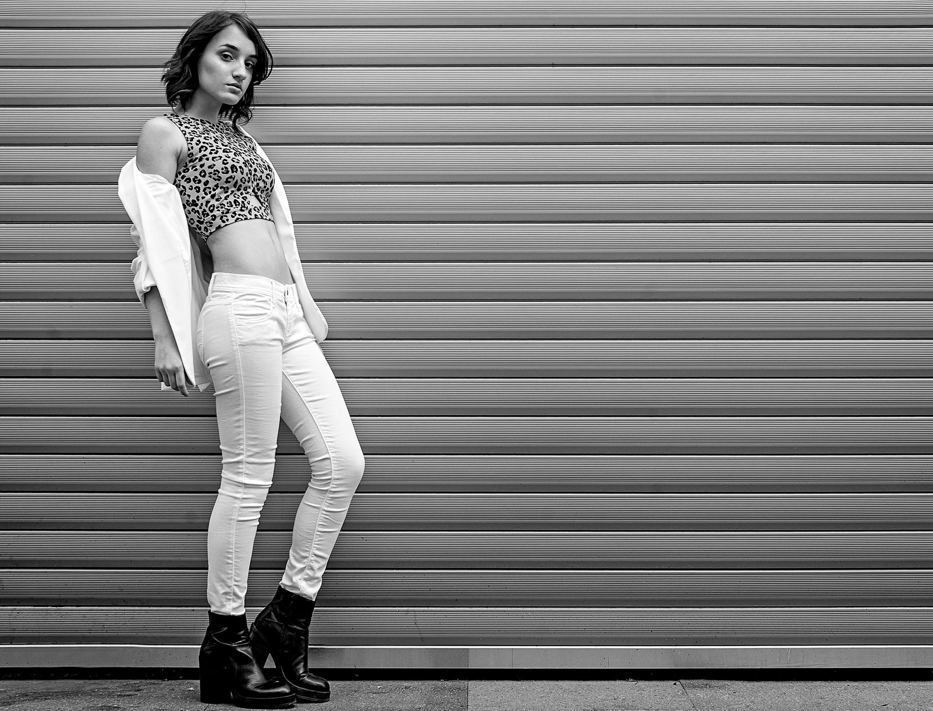 Una donna con un top leopardato e jeans bianchi posa vicino a una parete di metallo ondulato.