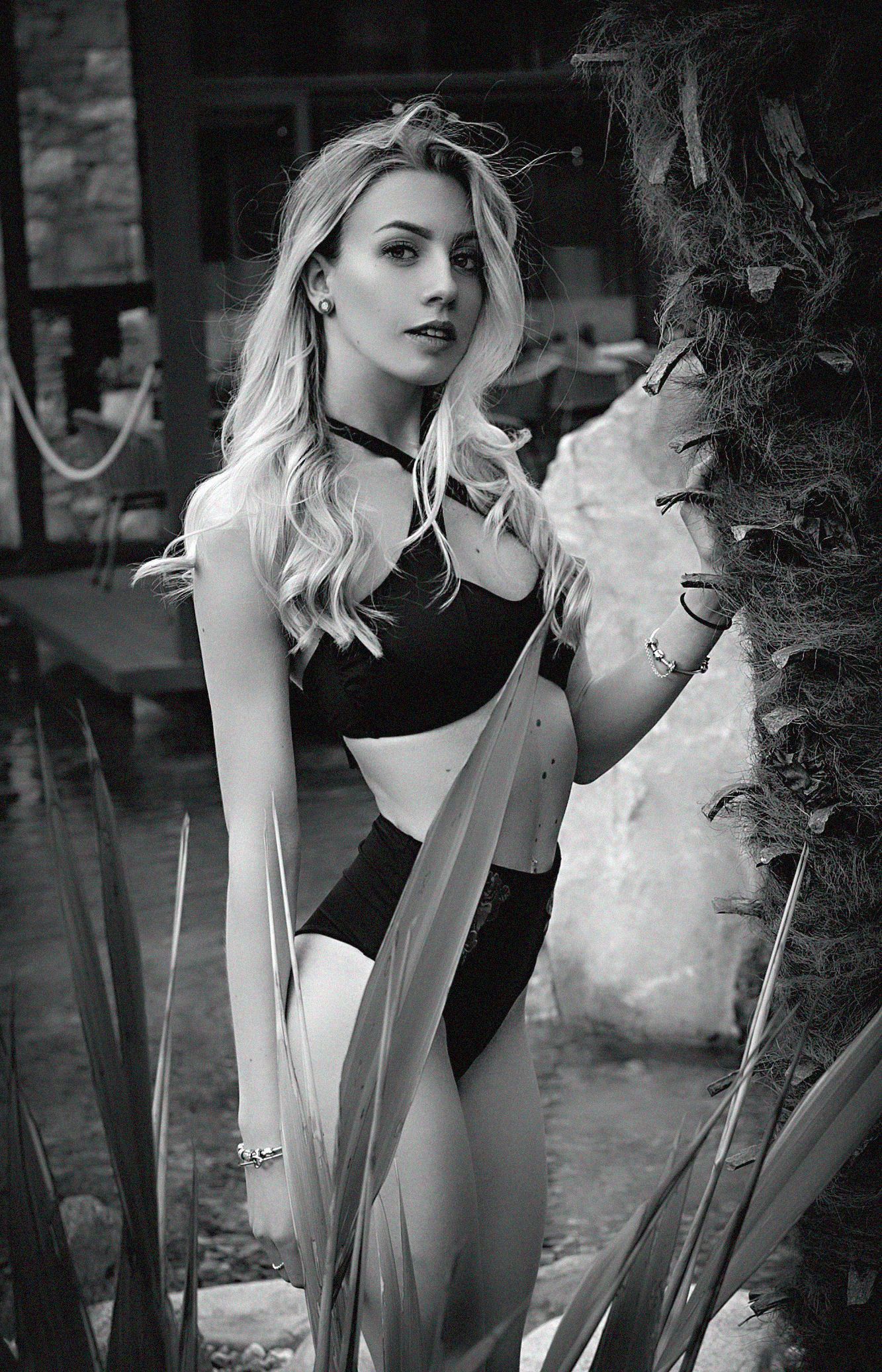 Una donna bionda in lingerie nera posa all'aperto vicino al fogliame.