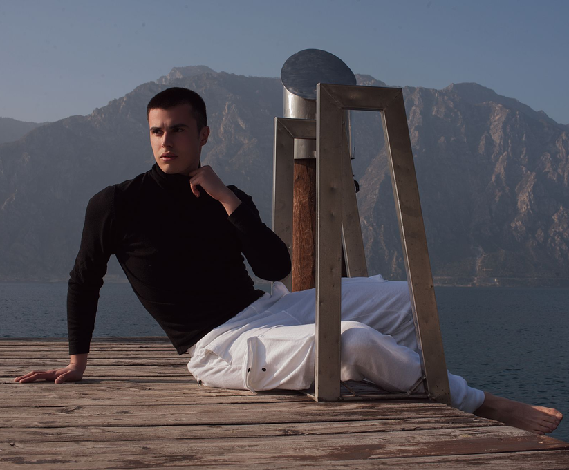 Un uomo con dolcevita nero e pantaloni bianchi posa su un molo, con montagne e acqua sullo sfondo.