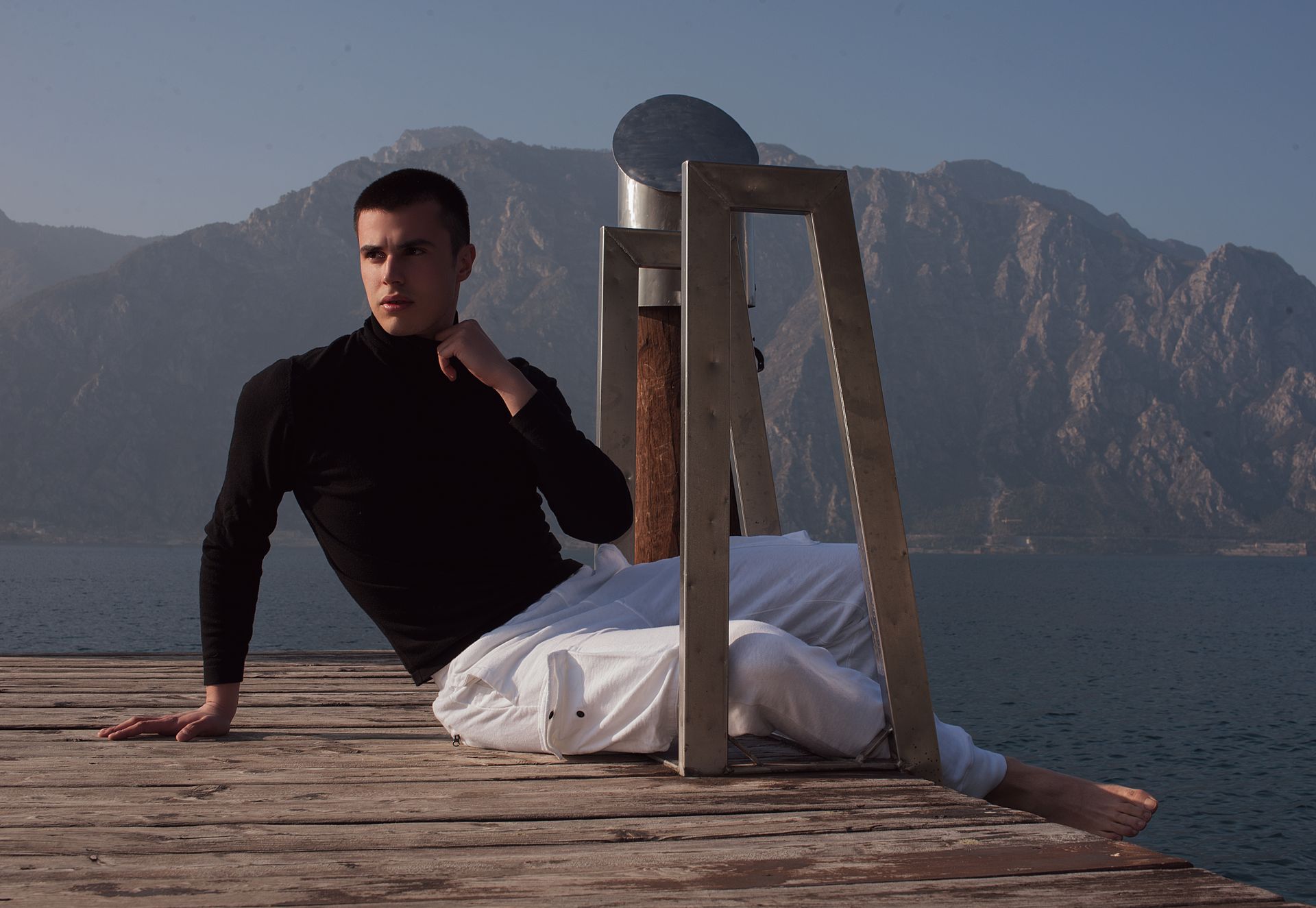 Un uomo con dolcevita nero e pantaloni bianchi posa su un molo di legno, con montagne e acqua sullo sfondo.