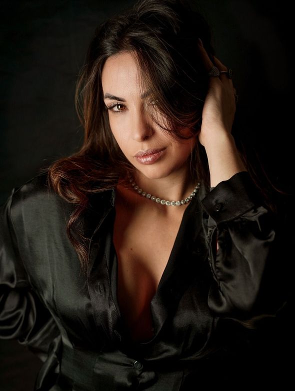 Donna con blazer di raso nero e collana di perle, guarda verso la telecamera con espressione contemplativa.