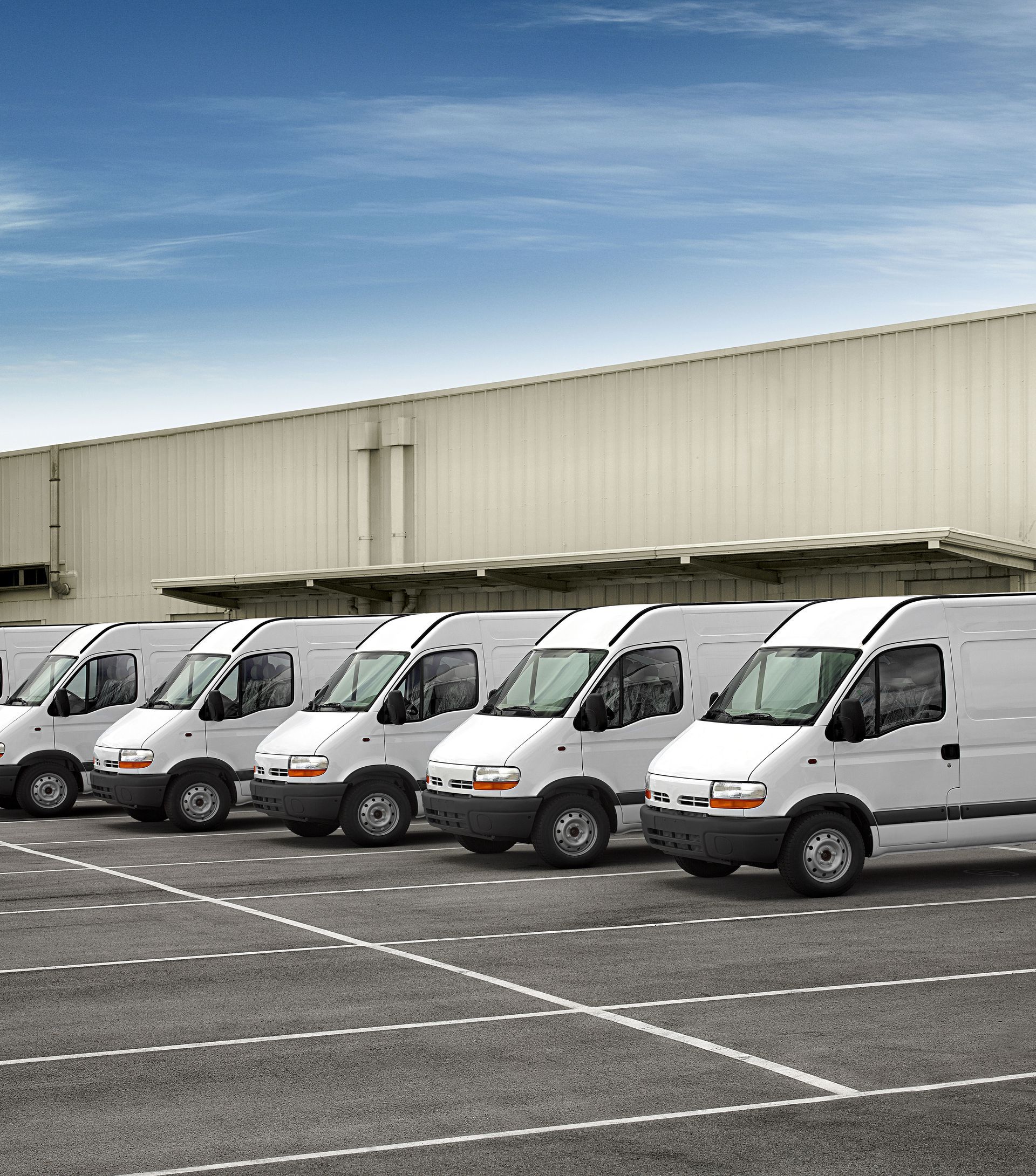 A row of white vans are parked in a parking lot in front of a building.