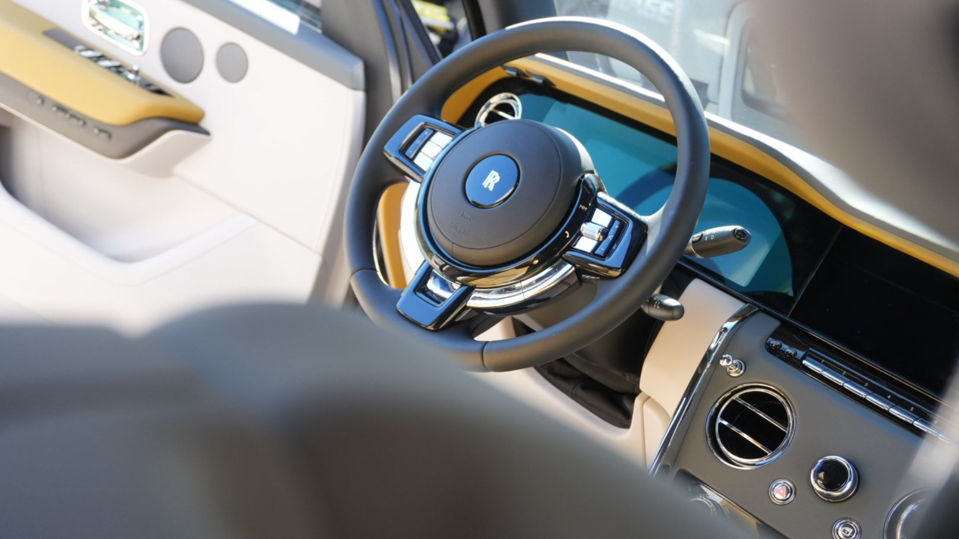 Interior view of a Rolls-Royce, focus on the steering wheel, tan leather, chrome accents, and dashboard.