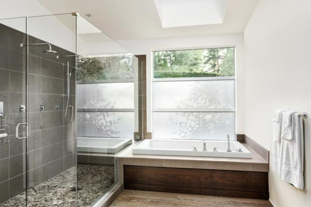 Modern bathroom with glass shower, stone floor, and white tub under a window.
