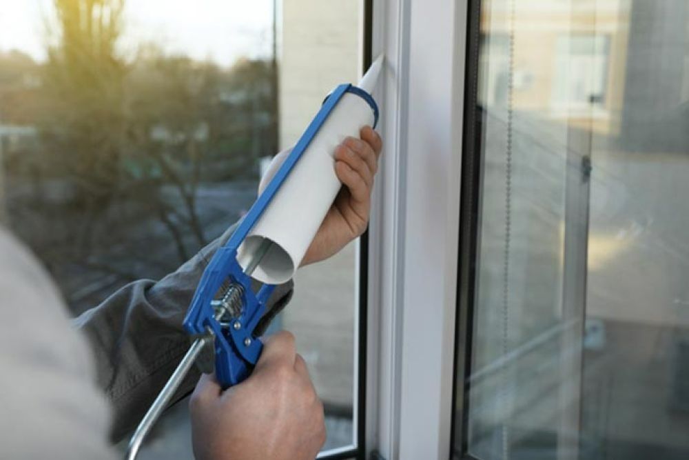 Person caulking a window frame with a caulk gun; white frame, blue gun, window reflection.