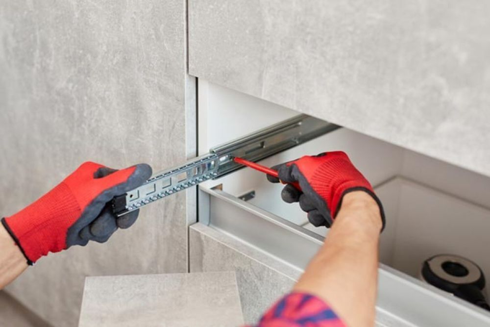 Person wearing red gloves, installing a drawer slide with a screwdriver.