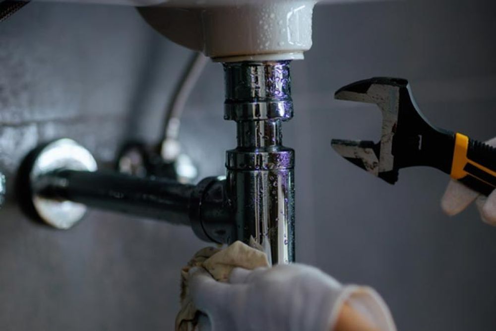 Plumber's hand with wrench near bathroom sink drain, with wet chrome pipes and rag.
